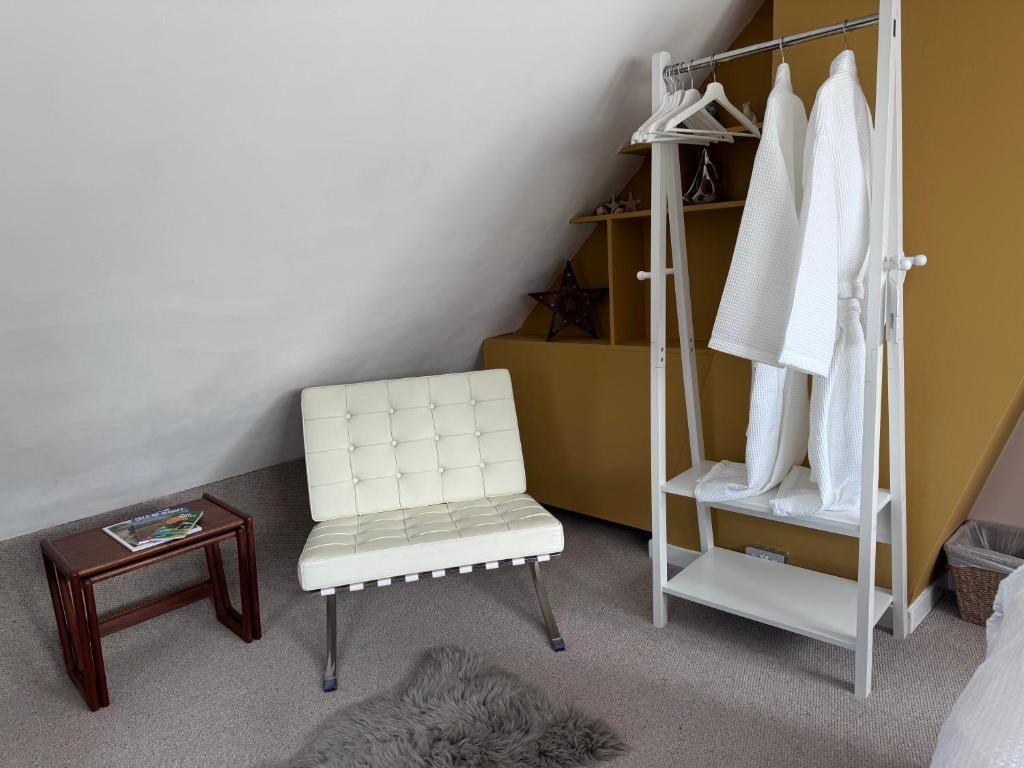 A room with a white tufted chair, a wooden side table with magazines, a white hanging clothes rack, and a grey furry rug.
