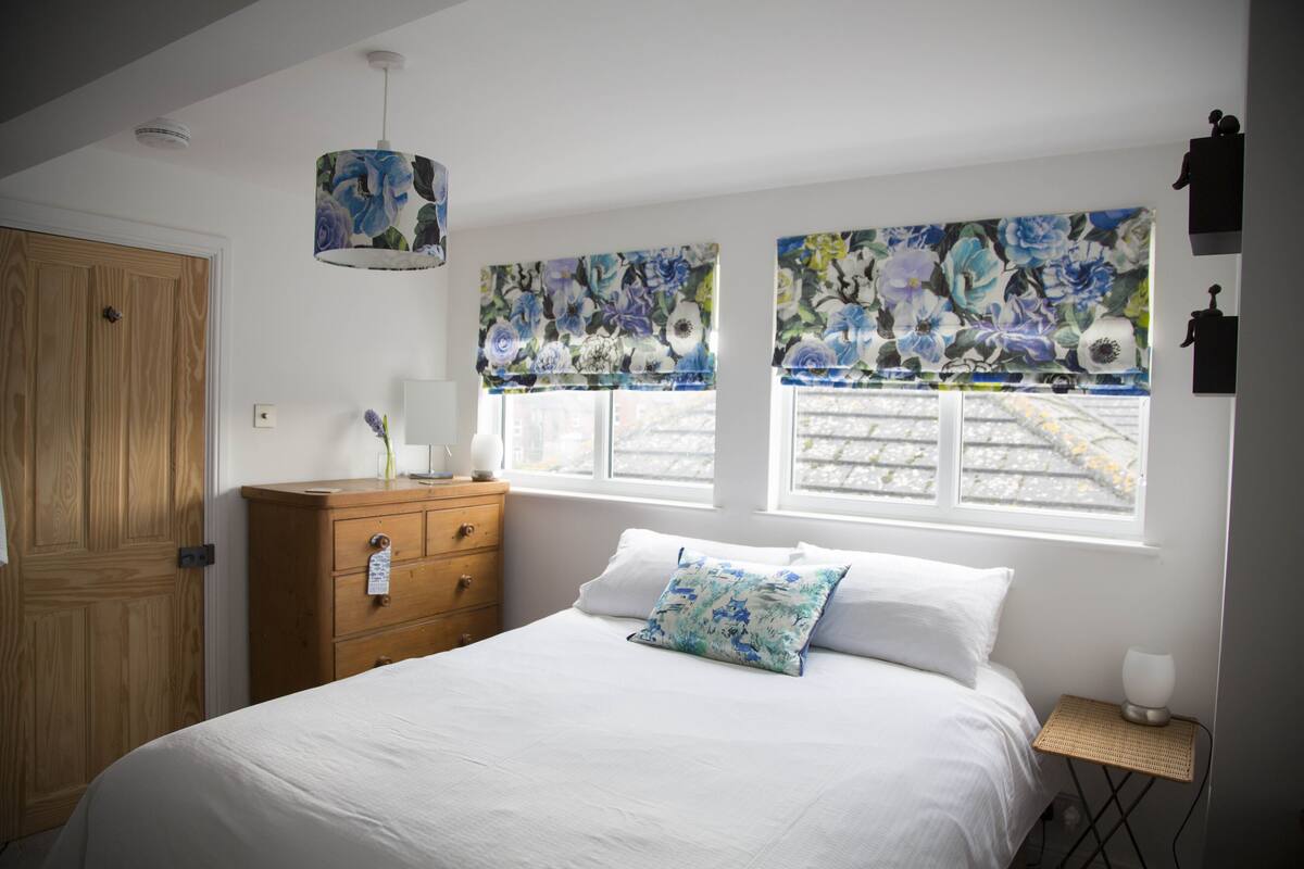 Une chambre avec une commode en bois, un lit avec une literie blanche, un coussin à motifs floraux et une fenêtre donnant sur la rue.