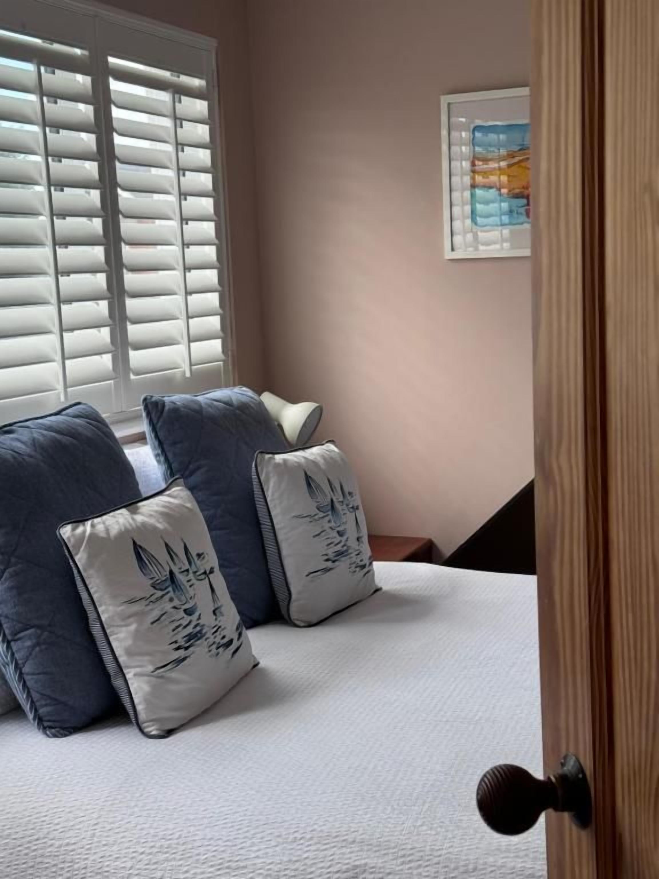 Quarto com cama, almofadas, janela com persianas, um quadro na parede e uma porta.