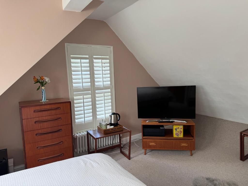 Un dormitorio con cama, cómoda, televisor sobre un soporte de madera, radiador y ventana con persianas.