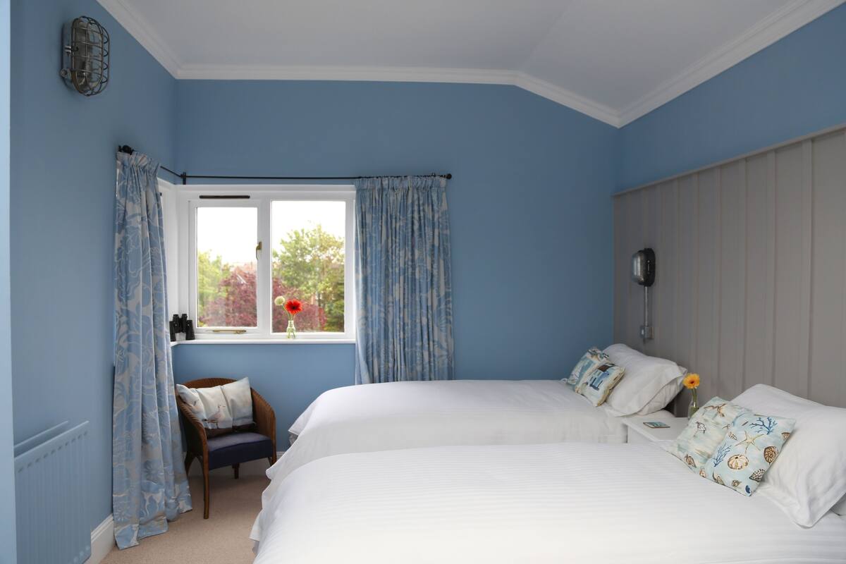 Une chambre avec deux lits, une fenêtre avec des rideaux, une chaise et une table de chevet ornée d’un vase de fleurs.