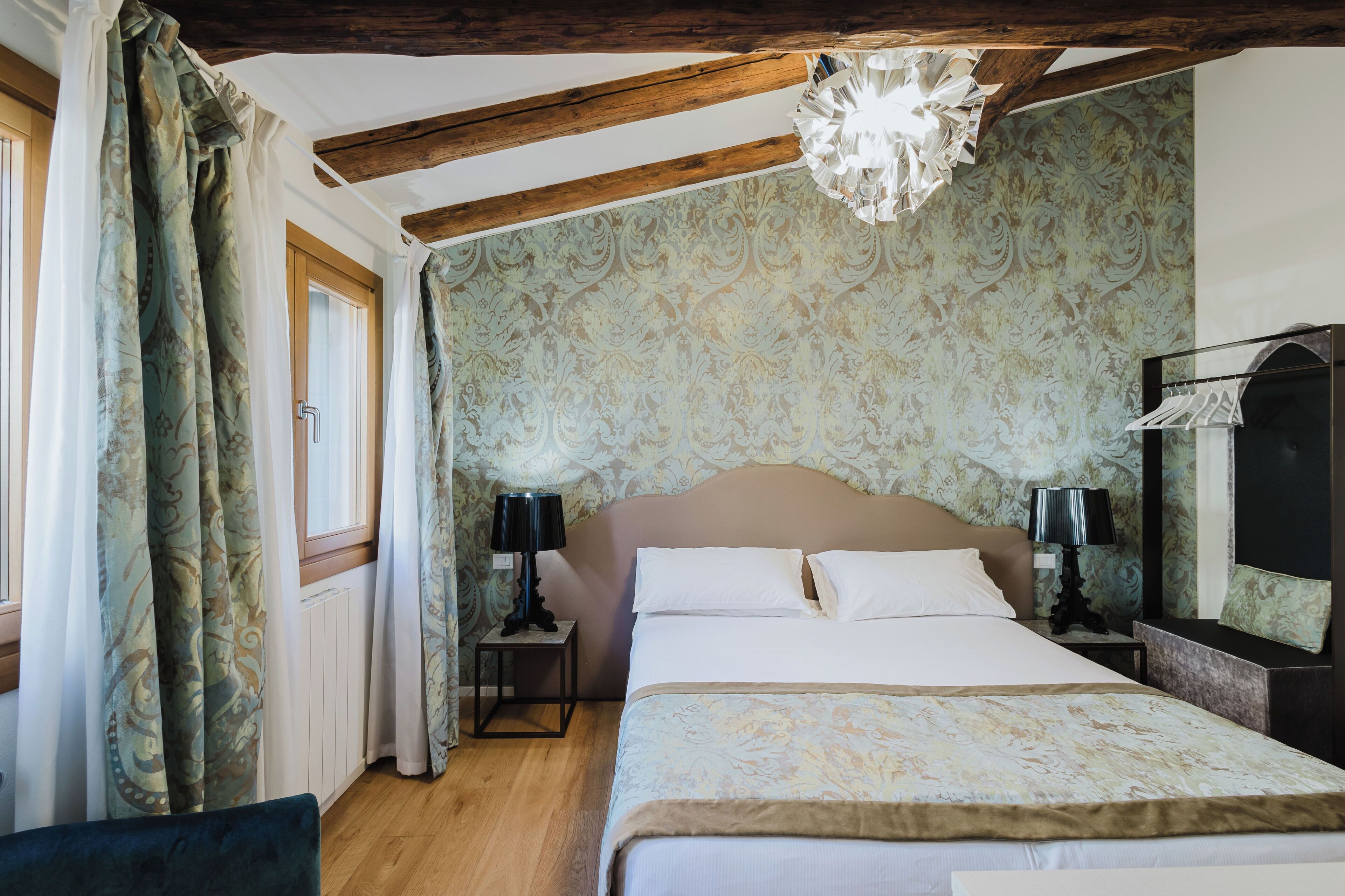 A bedroom with a large bed, two bedside tables with lamps, a wooden ceiling, patterned wallpaper, and a window with curtains.