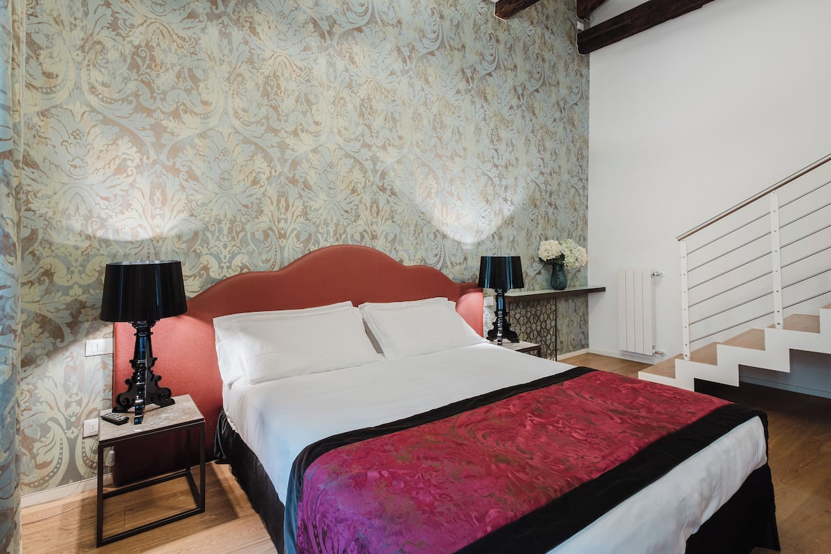 Una camera da letto con un letto grande, due comodini con lampade, una testiera rossa, un termosifone bianco e una scala.