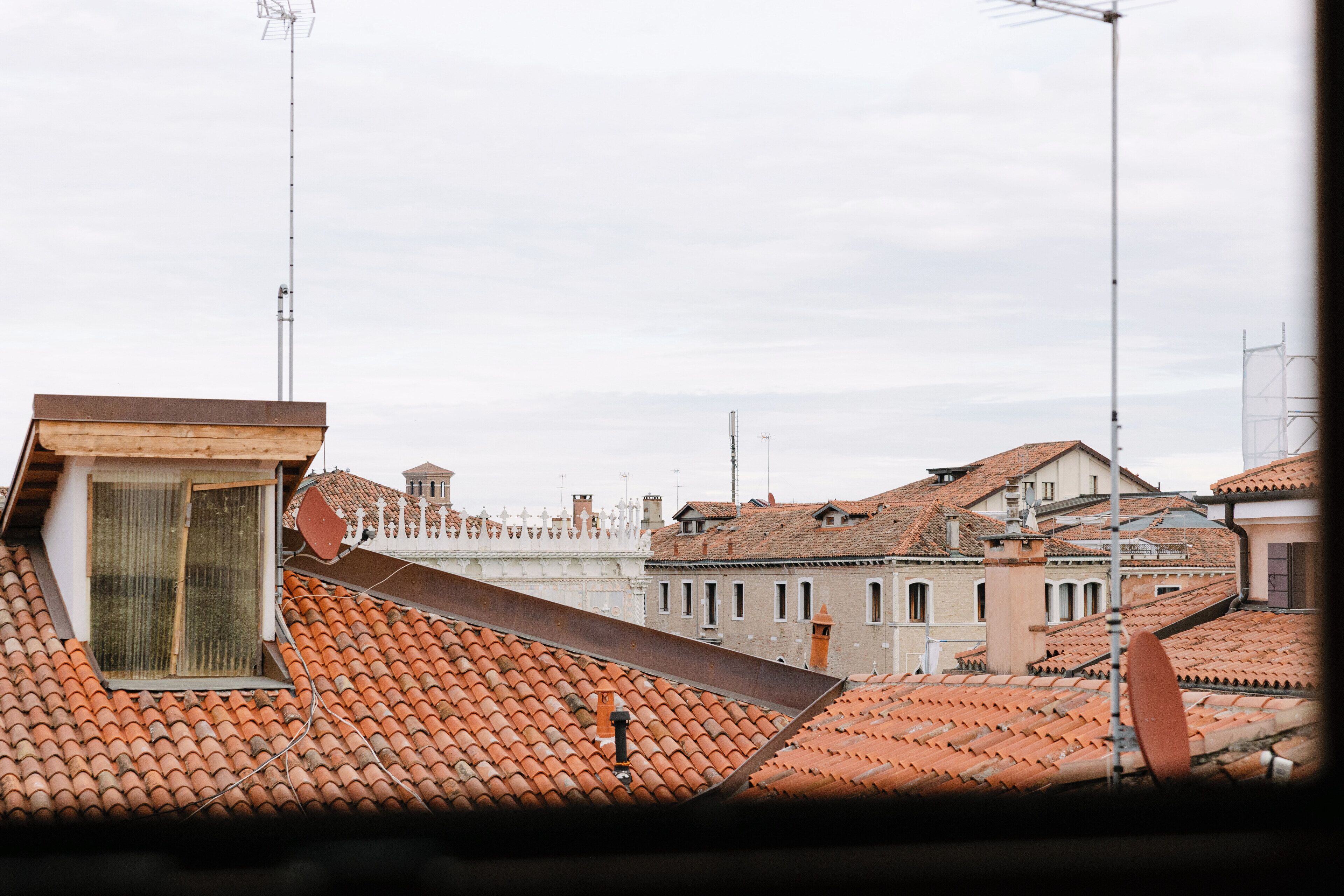 Blick auf Terrakotta-Dachziegel mit Satellitenschüsseln, ein kleines Fenster und ein weißes Gebäude mit Balkon im Hintergrund.
