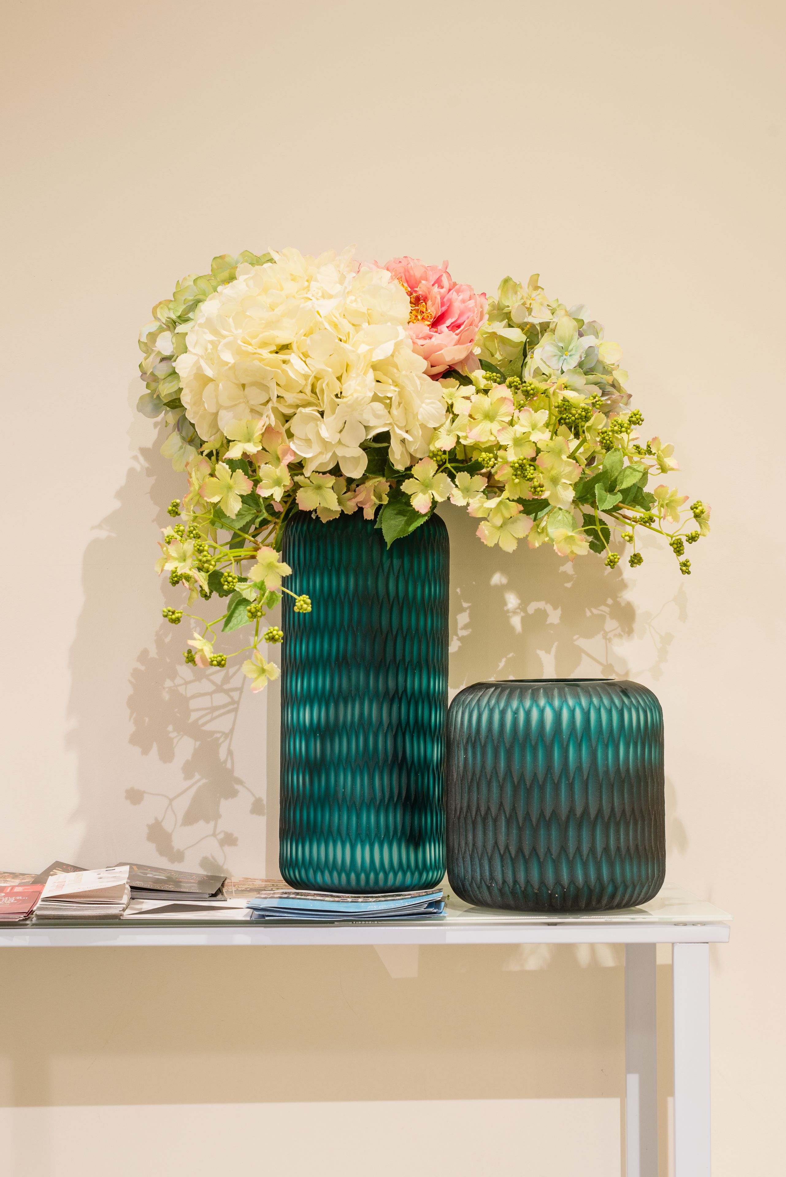 A teal vase with a floral arrangement on a white table against a plain wall.