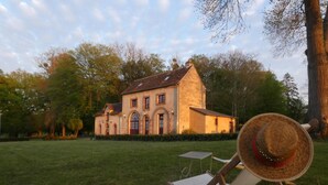 Un grand bâtiment en pierre, doté de fenêtres en arc de cercle et d’une cheminée, entouré d’une pelouse verdoyante et d’arbres.