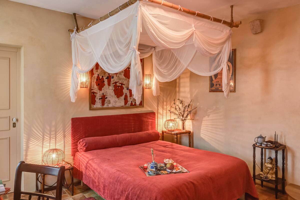 Een kamer met een bed, een stoel, een tafel met een Boeddhabeeld en een dienblad met eten en drinken.