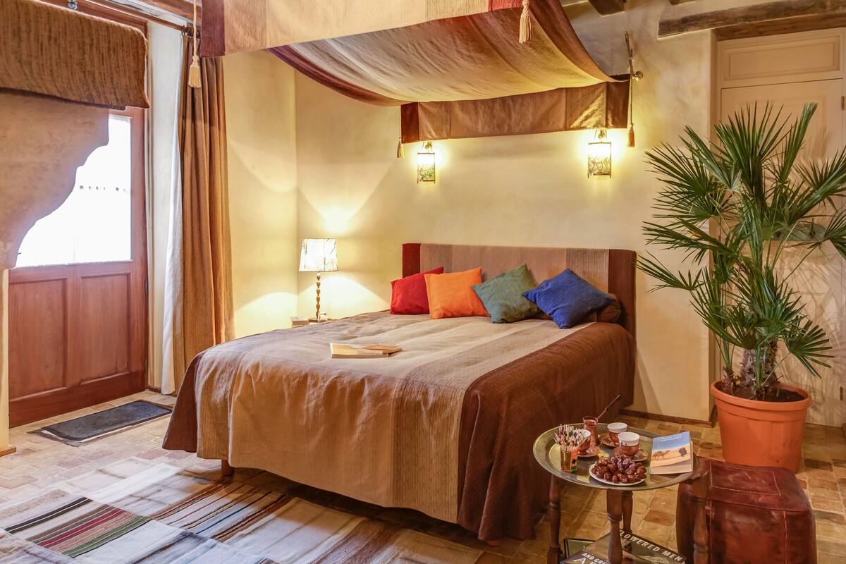 Een gezellige slaapkamer met een groot bed, een nachtkastje met een lamp, een kamerplant en uitzicht naar buiten.