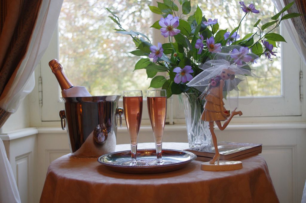 Uma mesa com uma garrafa de champanhe, duas taças, um vaso com flores e uma figurinha.