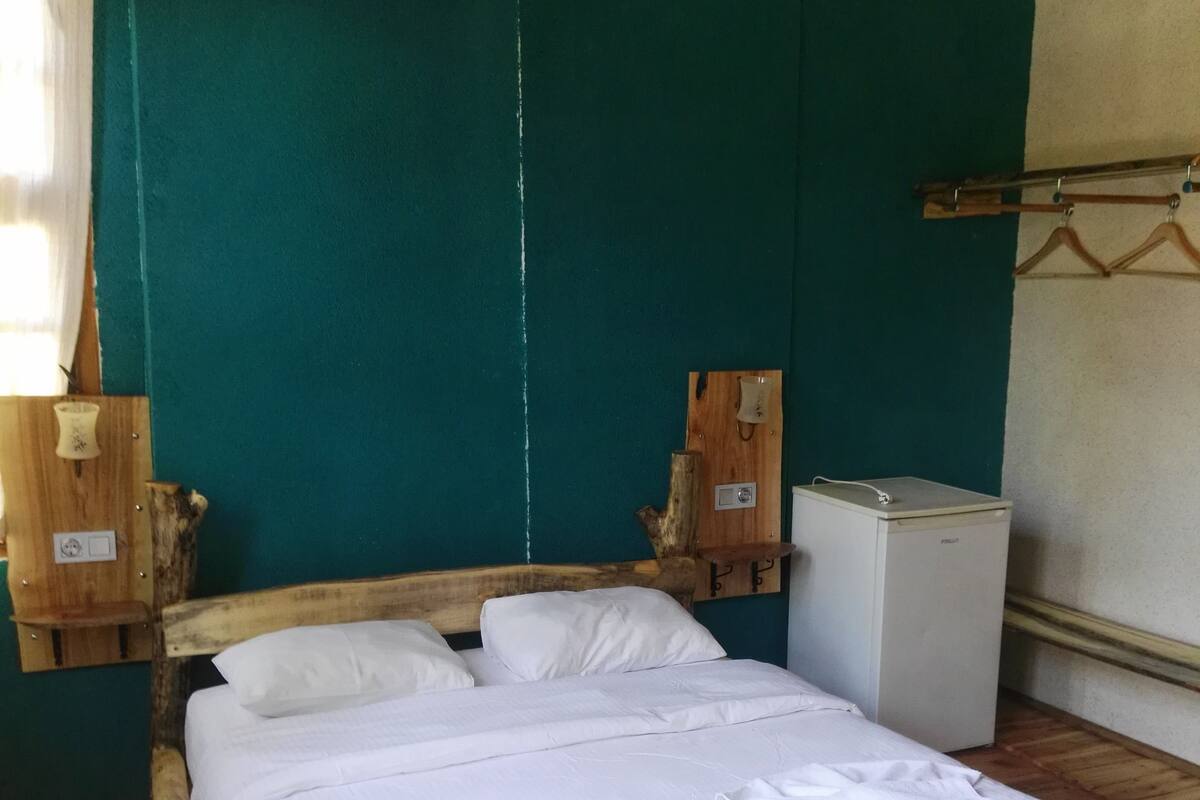 A wooden bed with white bedding and towels, a small white refrigerator, and a teal wall.
