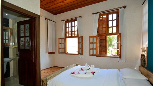 Una habitación con una cama de madera, dos ventanas con cortinas, techo de madera y una puerta de madera que da a un baño.