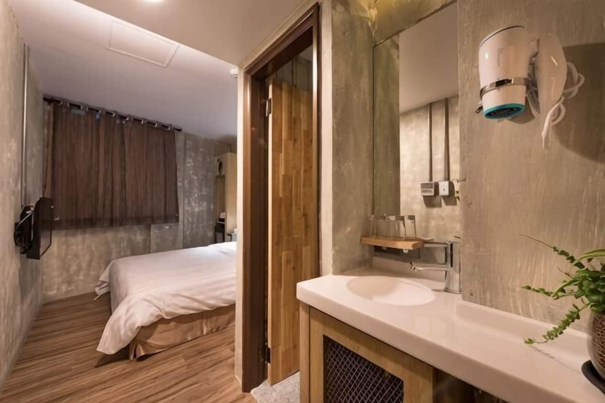 A hotel room with a wooden floor, a bed, a bathroom with a sink and mirror, and a shower area.