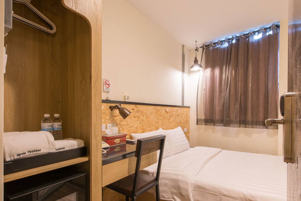 A hotel room with a bed, a desk with a chair, a TV, and a window with curtains.