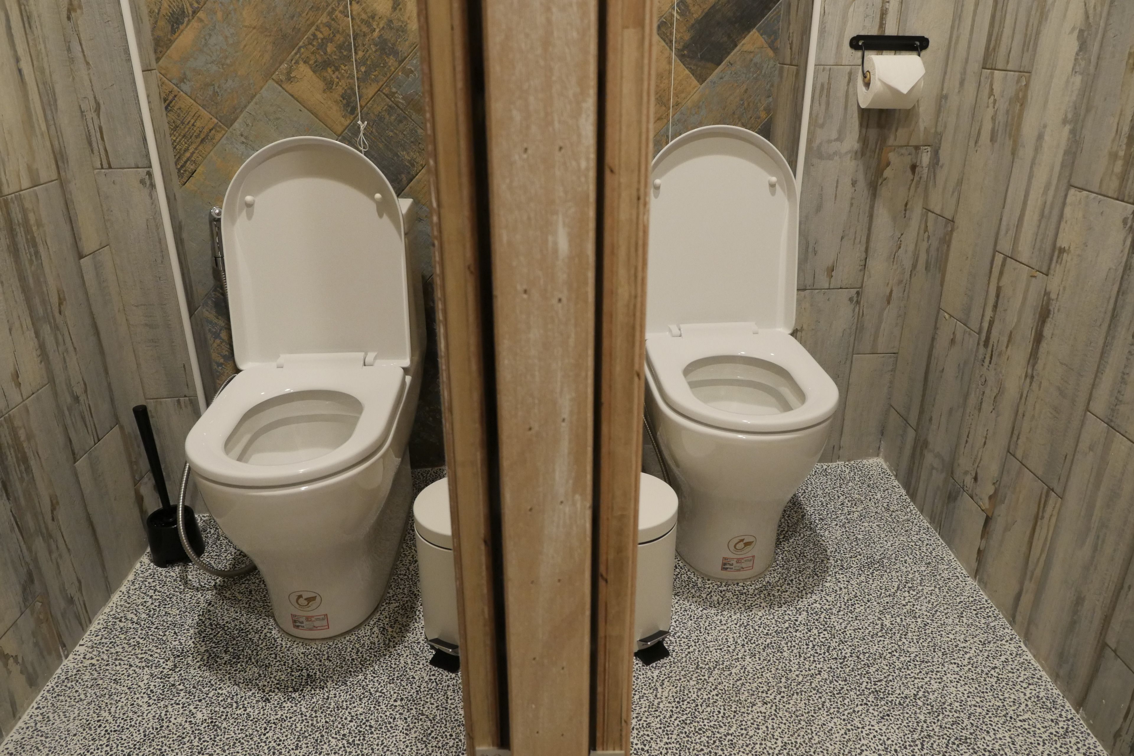 A bathroom with two toilets, a toilet brush, and a toilet paper holder on the wall.