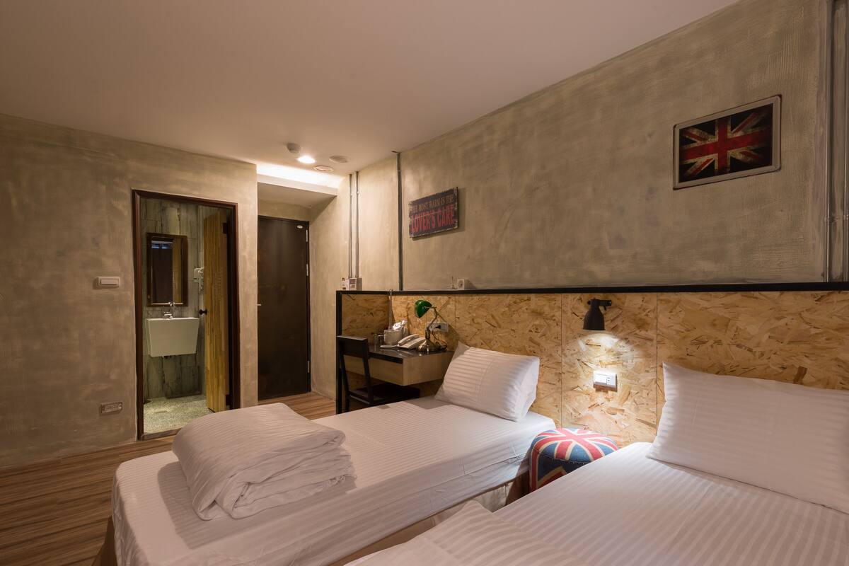 A hotel room with two beds, a wooden headboard, a small desk with a lamp, a mirror, and a door leading to a bathroom.