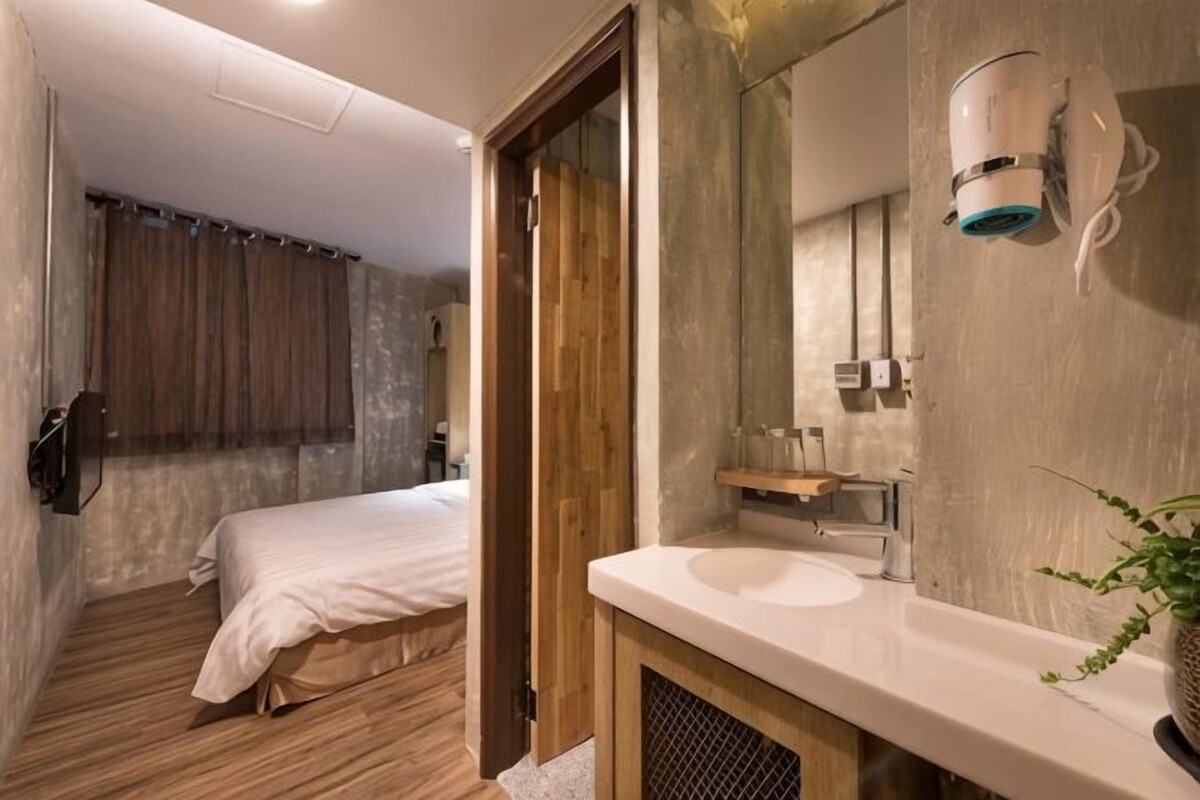 A hotel room with a wooden floor, a bed, a bathroom with a sink and mirror, and a shower area.