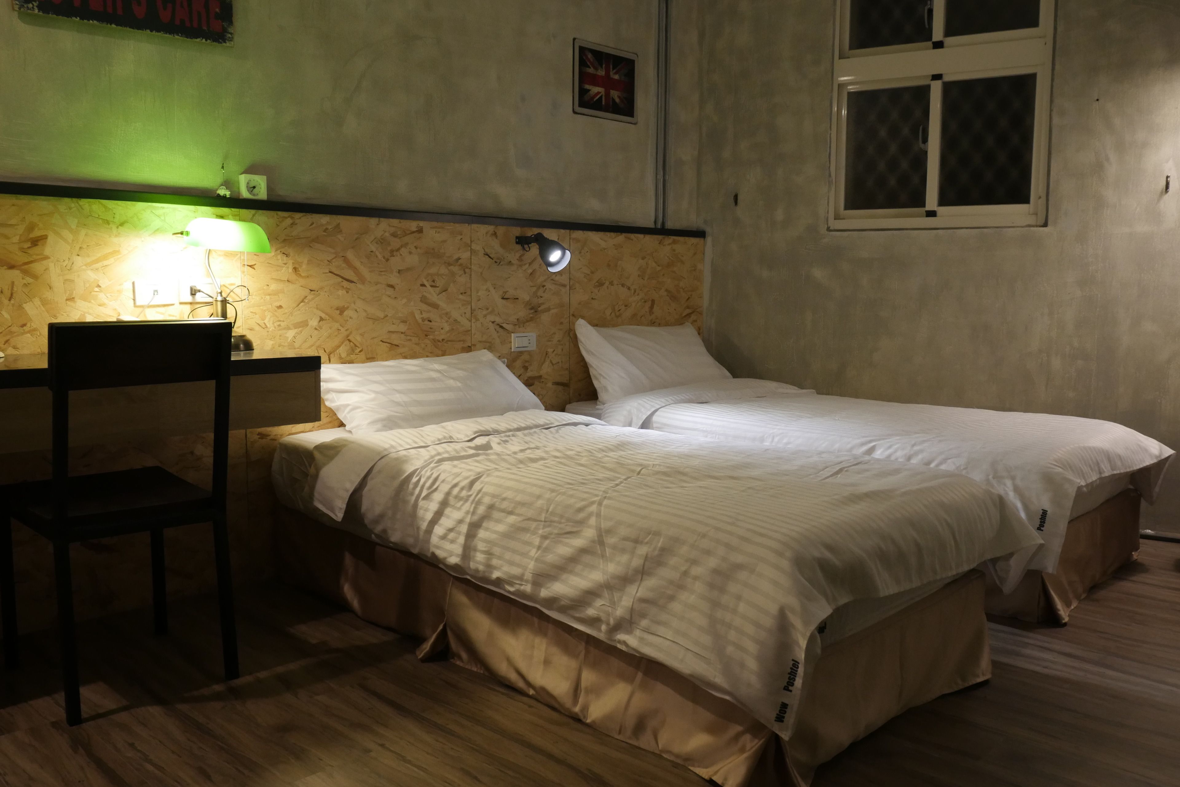 A room with two single beds, a desk with a chair, a green desk lamp, and a small flag on the wall.