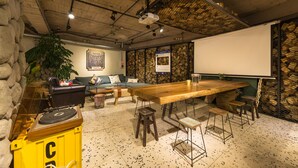 A room with a wooden table, bar stools, a projector screen, a sofa, and a large plant.