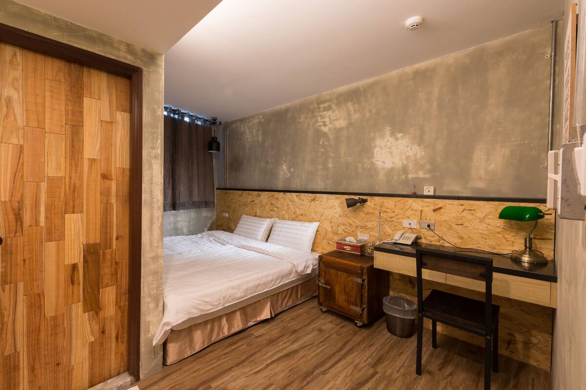 A compact room with a wooden door, a bed, a desk with a lamp, and a small trash bin.