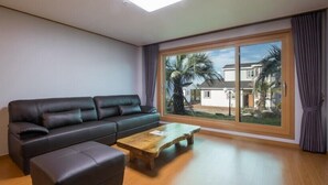 A modern living room with a black leather sofa, a wooden coffee table, and a large window with a view of palm trees and houses. - The Some Pension (Seogwipo)