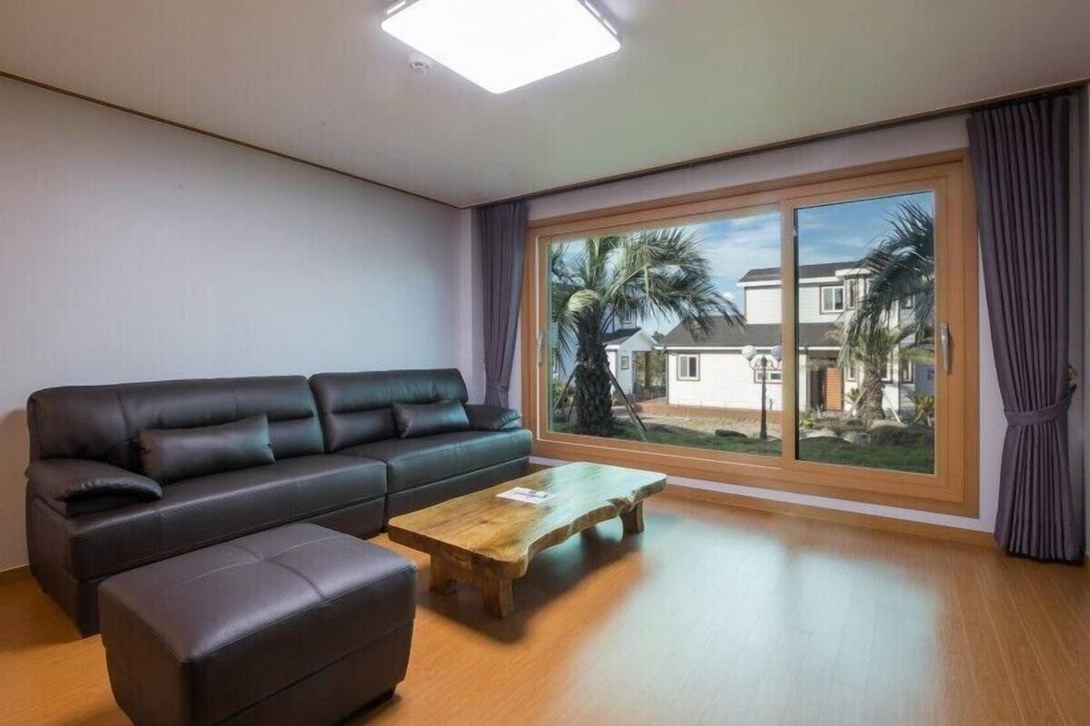 A modern living room with a black leather sofa, a wooden coffee table, and a large window with a view of palm trees and houses.