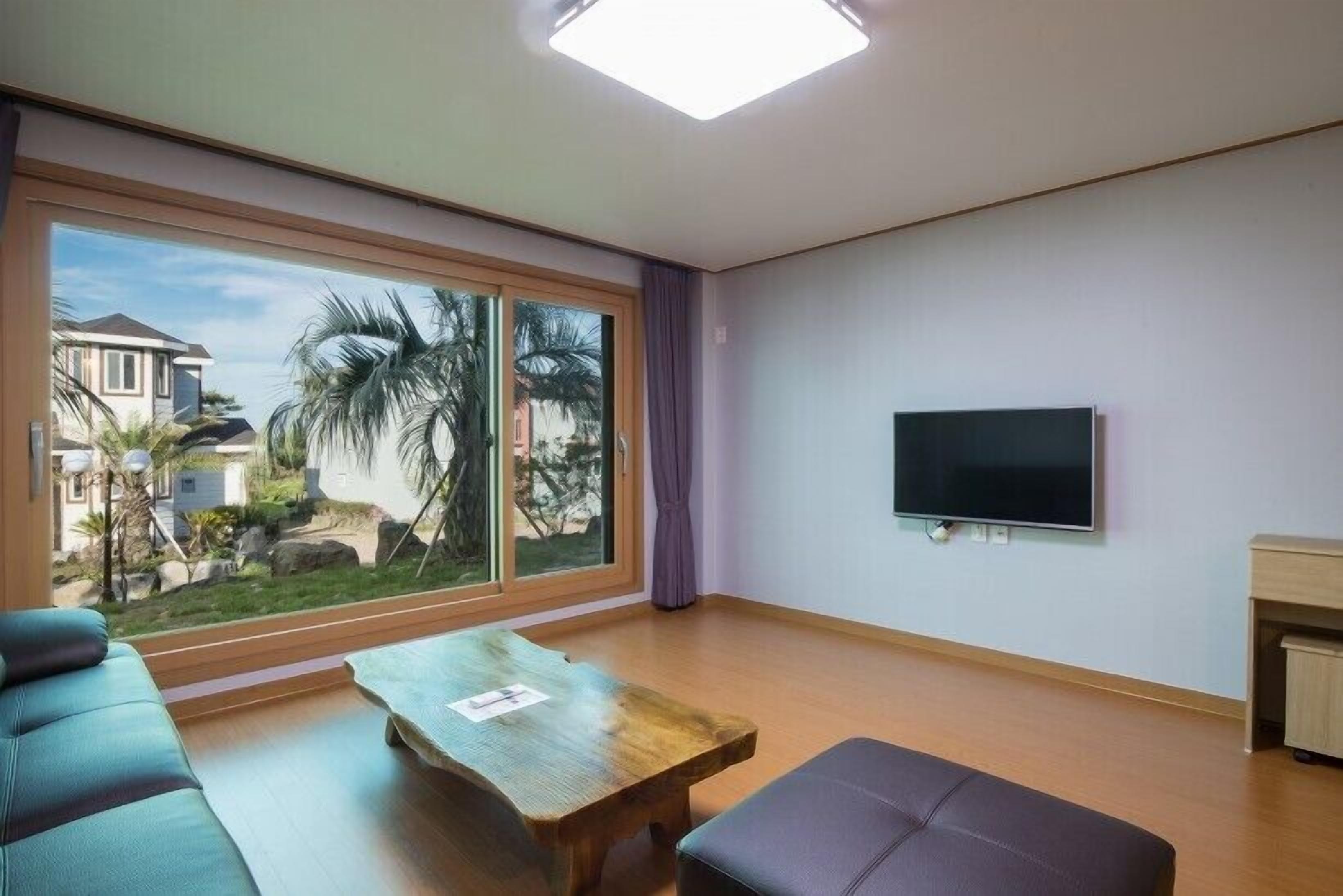 A modern living room with a large window, a flat-screen TV, a wooden coffee table, a sofa, and a small cabinet.