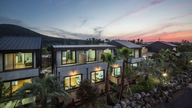 A modern multi-story residential building with large windows, a well-lit interior, and a landscaped garden with palm trees. - The Some Pension (Seogwipo)