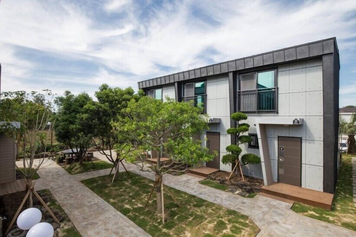 A modern two-story building with a flat roof, large windows, and a well-maintained garden with trees and a stone pathway.