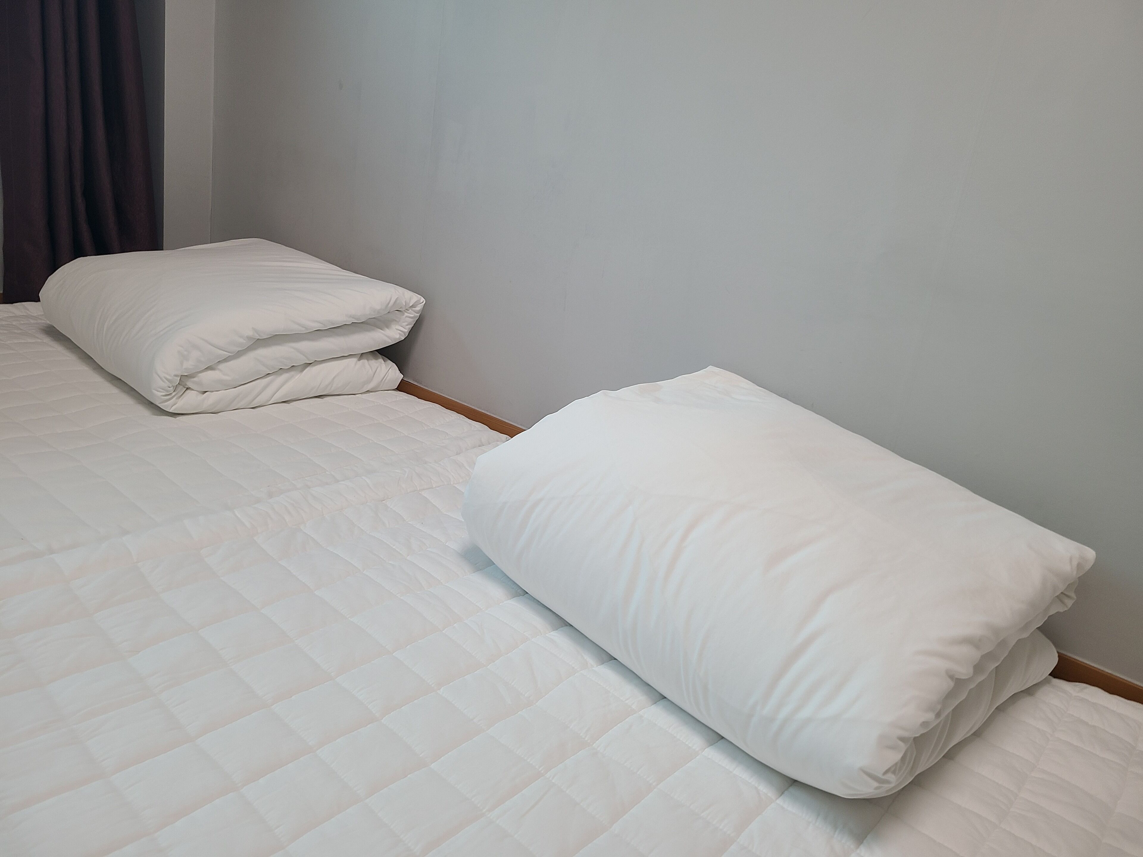 A neatly made bed with white bedding in a simple room with a plain wall and a dark curtain.