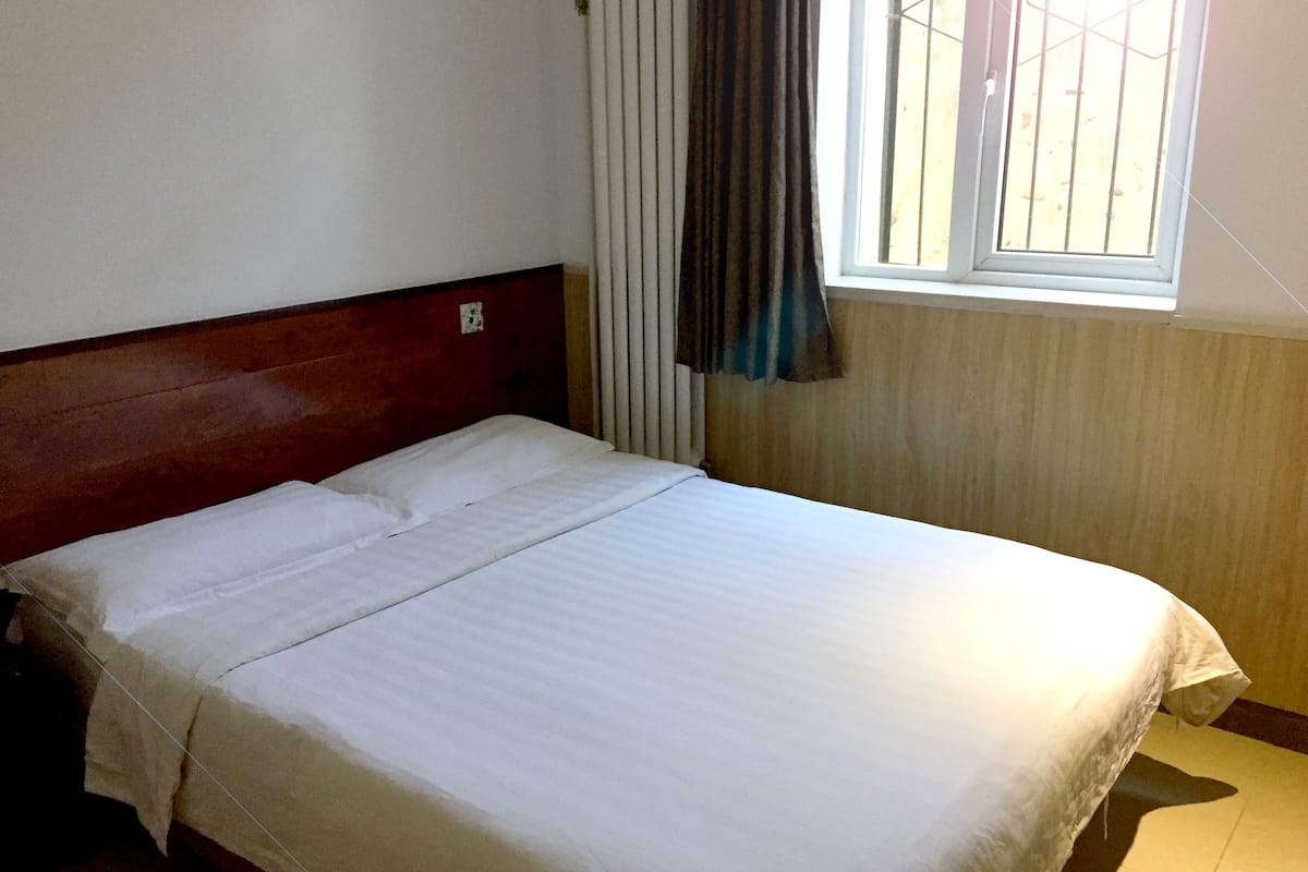Una cama bien tendida en una habitación con cabecera de madera, una ventana con persianas y un radiador.