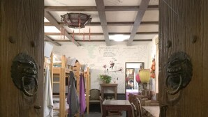 A room with a high ceiling, exposed beams, a wooden table, chairs, and a desk with a chair. There are shelves with items and a hanging lantern. - 365 inn - Hostel (Beijing)