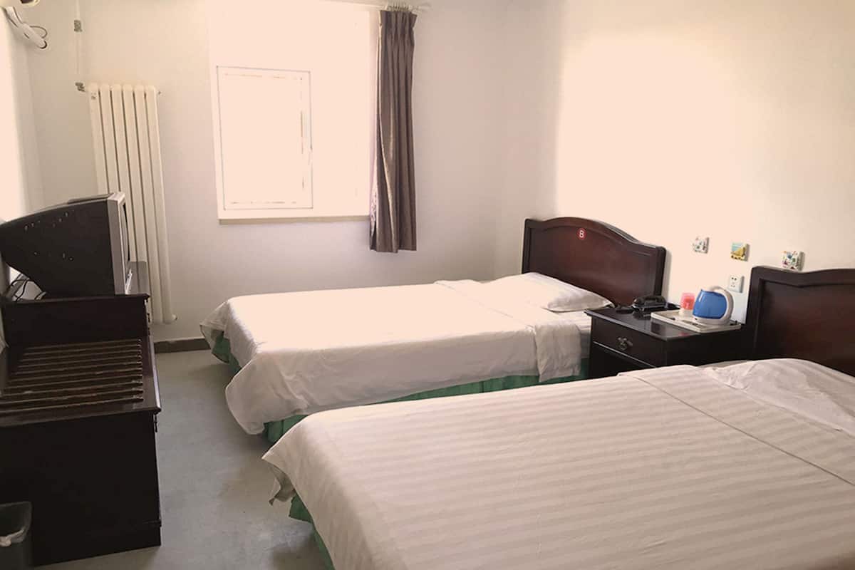 Habitación de hotel con dos camas individuales, cómoda de madera, escritorio con silla, radiador y ventana con cortinas.