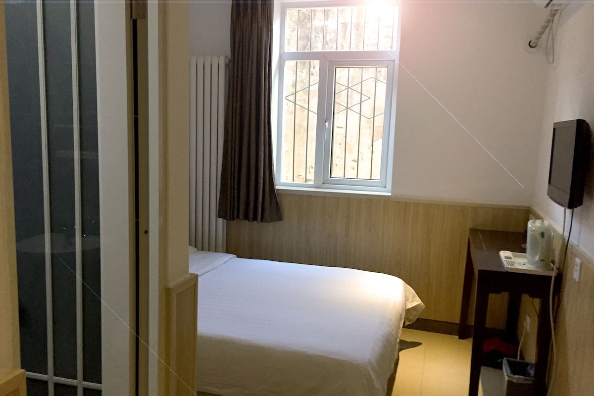 Un dormitorio pequeño con una cama individual, una ventana con cortinas, una mesita de noche con lámpara y un televisor montado en la pared.