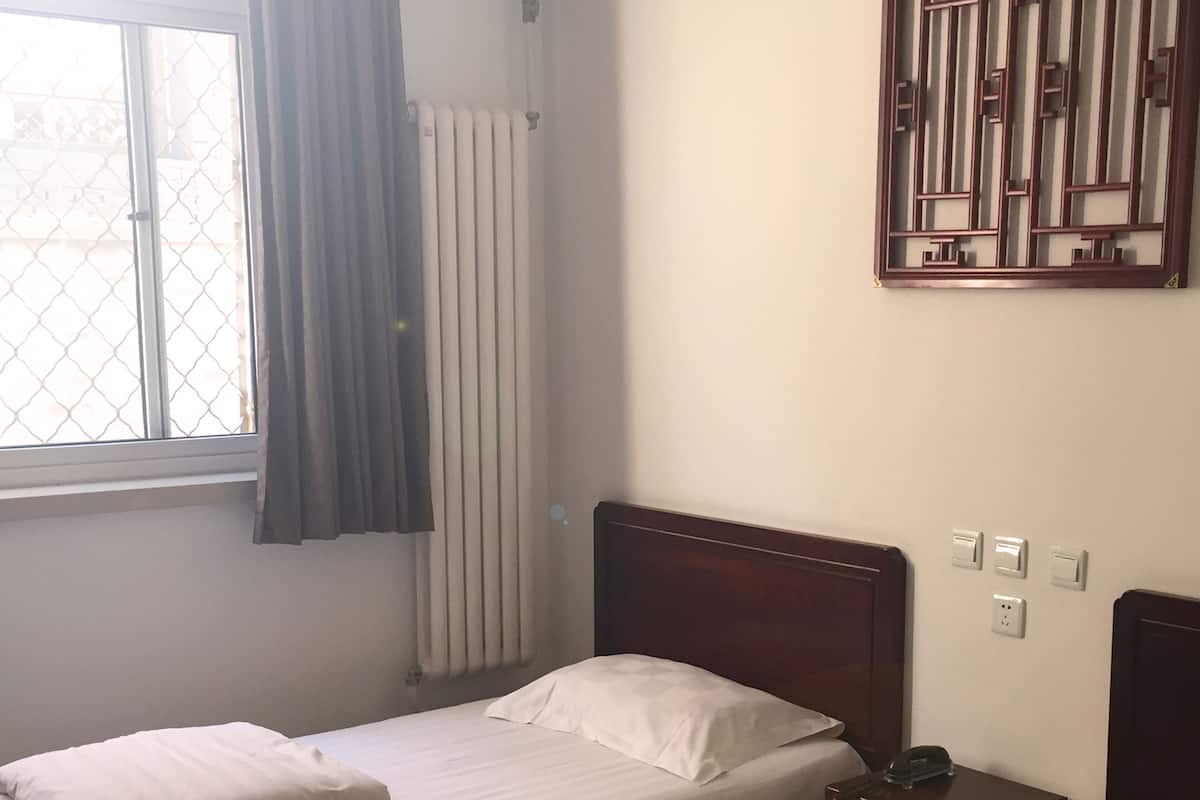 Una habitación con dos camas individuales, un radiador, una ventana con cortinas, un panel decorativo en la pared y una mesita de noche con lámpara.