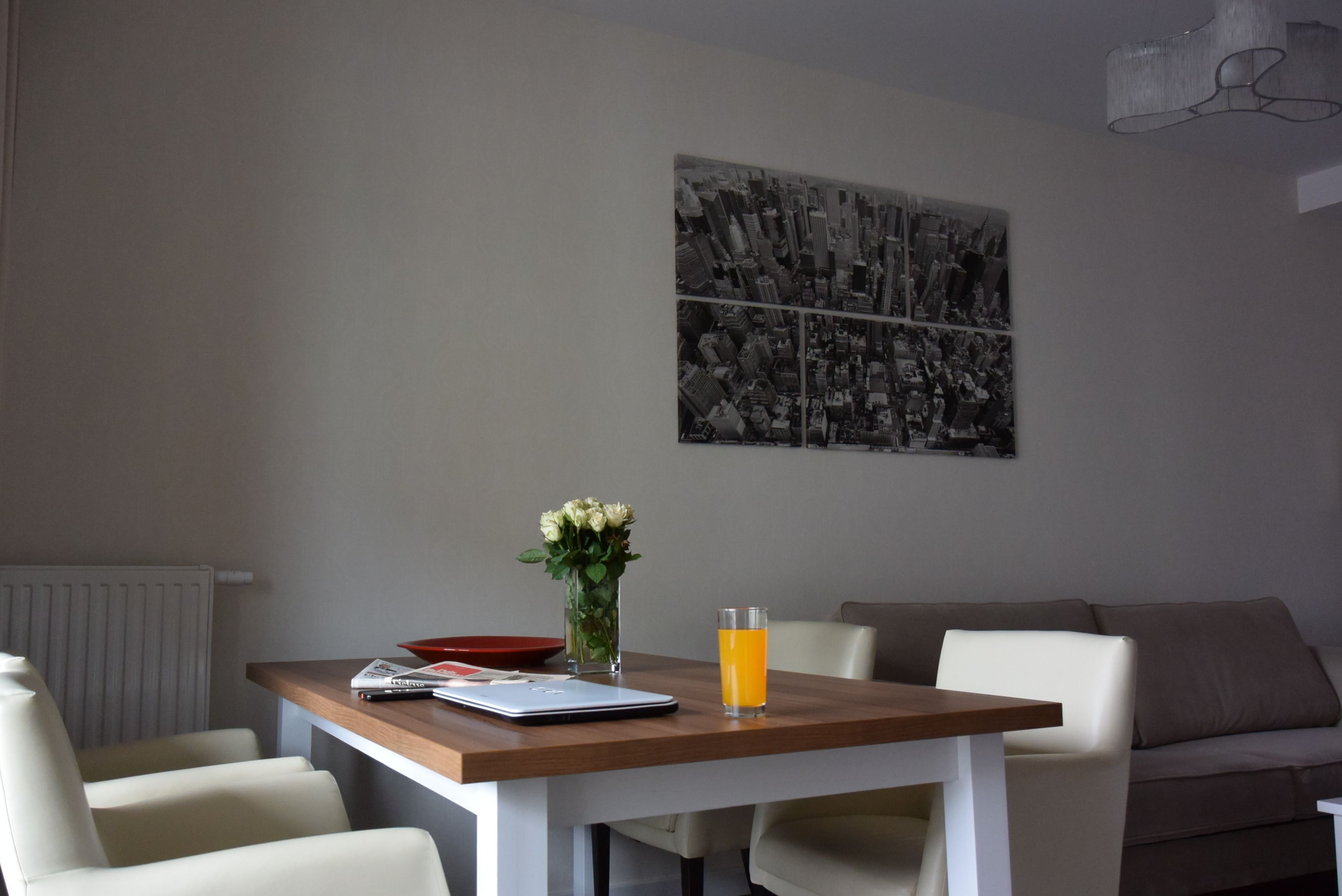 Un comedor con una mesa, sillas, un florero con flores, un vaso de jugo de naranja y un cuadro de paisaje urbano en blanco y negro en la pared.