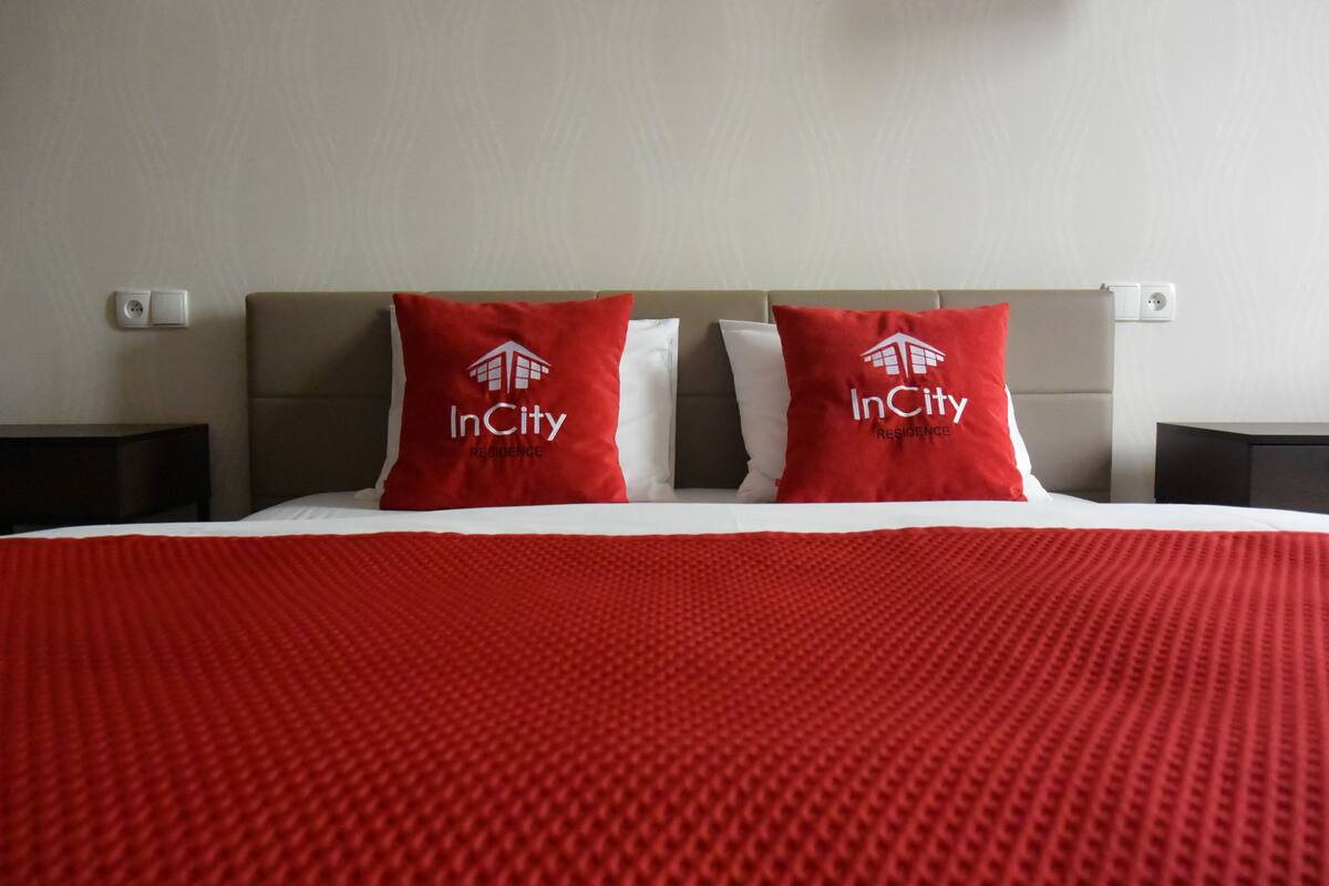 Quarto de hotel com uma cama, duas almofadas vermelhas com o logotipo InCity, um cobertor vermelho texturizado e uma cabeceira cinza.