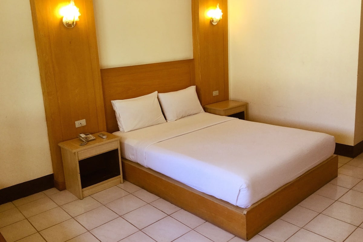 A standard hotel room with a wooden headboard, two bedside lamps, a bed with white linens, a nightstand with a telephone, and a small shelf.