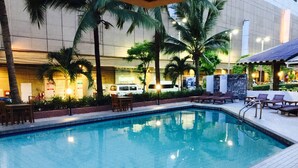 Une piscine entourée de chaises longues et de palmiers, le tout bordé par un bâtiment à la façade recouverte de carreaux.