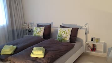 Una cama bien hecha con un edredón marrón y dos toallas verdes, una mesita de noche con una lámpara, y un estante pequeño con libros y una vela.
