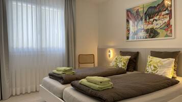 Quarto de hotel moderno com duas camas, uma janela grande com cortinas leves, um quadro pendurado na parede e uma pequena mesa com uma cadeira.