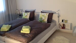 Een netjes opgemaakt bed met bruin beddengoed en twee groene kussens, een nachtkastje met een lamp en een klein plankje met boeken en een glas.