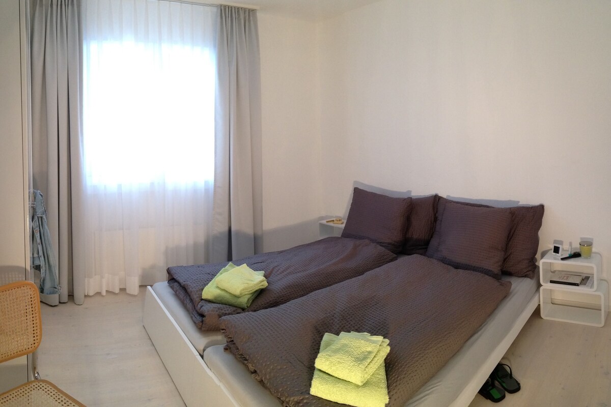 Quarto de hotel com cama, poltrona, mesa de cabeceira e janela com cortinas.
