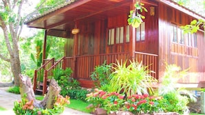 Een houten hut met een veranda, omgeven door weelderige begroeiing en bloeiende planten.
