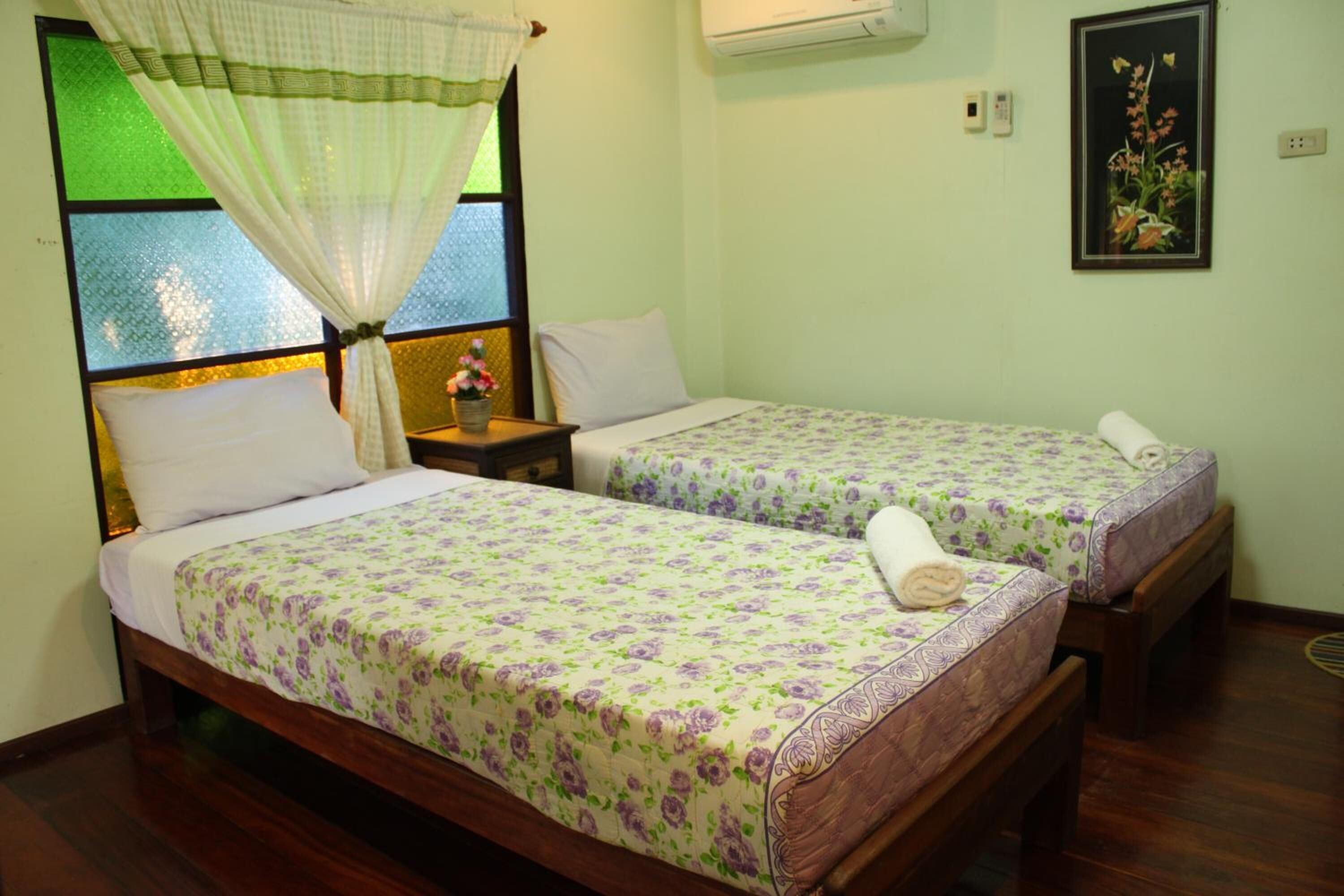 A room with two beds, each with floral bedding, a nightstand with a lamp, and a window with a green-tinted glass.