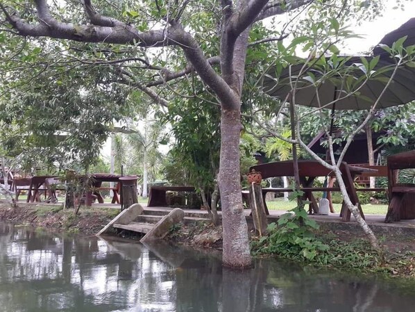 A serene outdoor area with a pond, wooden benches, and a covered seating area surrounded by lush greenery. - Baansuanlynjee Maeklong Resort (Bang Khonthi)