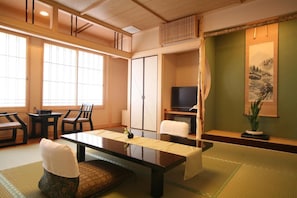 Een traditionele Japanse kamer met tatami vloerbedekking, een lage tafel, stoelen, een televisietoestel en een schilderij aan de muur.