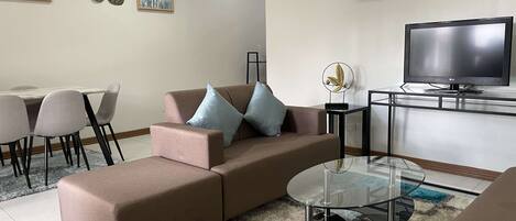 A modern living room with a brown sofa, two side tables, a glass coffee table, a flat-screen TV, and artwork on the walls.