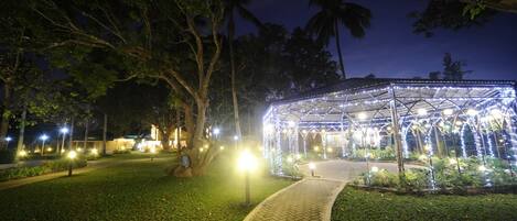 A well-lit gazebo with a brick pathway leading to it, surrounded by trees and illuminated by street lamps.