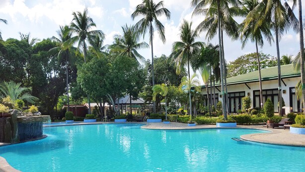 A swimming pool surrounded by palm trees and a building with a green roof. - Batangas Country Club (Batangas City)