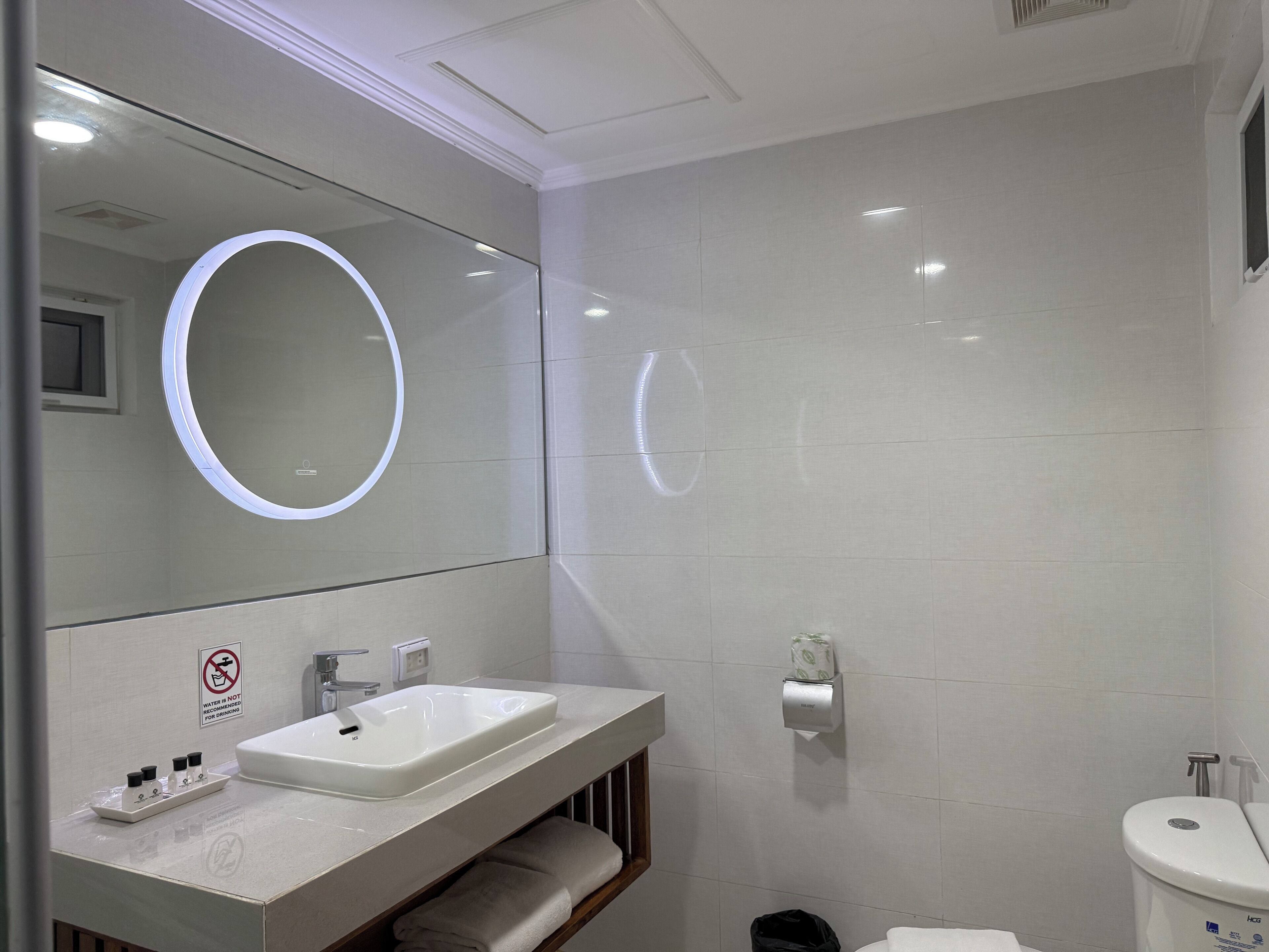 Uma casa de banho moderna com lavatório branco, espelho e sanita. Há um sinal de proibição de água na parede, um porta-toalhas e um cesto de lixo.